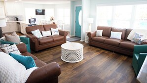 Een ruime woonkamer met twee bruine leren banken, een witte salontafel en teal fauteuils. In de achtergrond is een keuken zichtbaar met witte kasten en een flatscreen-tv.