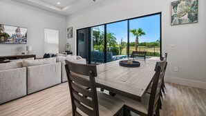 A spacious living room with a large dining table, comfortable seating, a ceiling fan, and a large window offering a view of the outdoors. - Liberty Grandview: Private Pool, Water Park, Golf Views (Kissimmee)