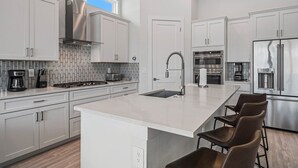 A modern kitchen with white cabinetry, a large island with bar stools, stainless steel appliances, and a tiled backsplash. - Liberty Grandview: Private Pool, Water Park, Golf Views (Kissimmee)