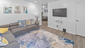A modern living room with a gray sofa, colorful throw pillows, a large abstract rug, and a flat-screen TV mounted on the wall.