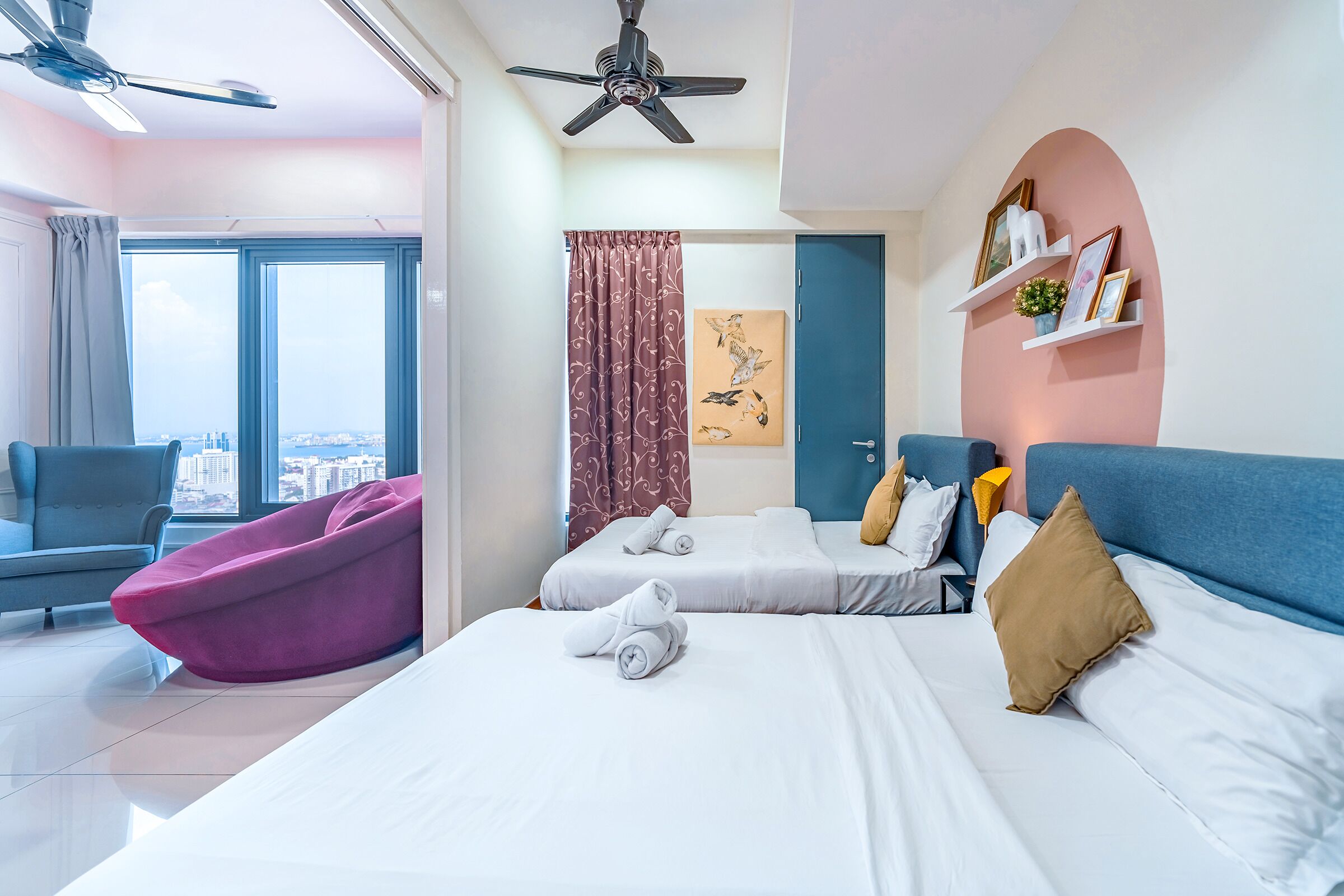 Habitación de hotel moderna con dos camas, un sillón informal tipo bean bag color rosa, vistas a la ciudad y un ventilador de techo.