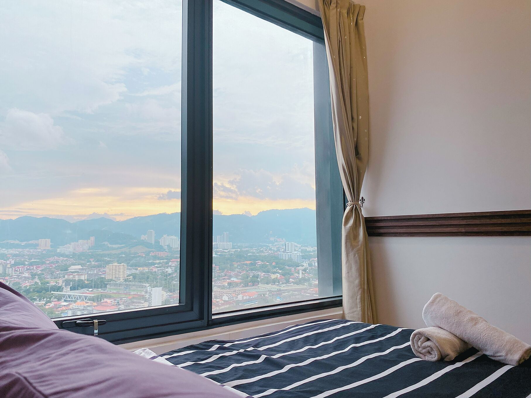 A hotel room with a large window offering a cityscape view, a bed with striped bedding, and a rolled towel on the bed.