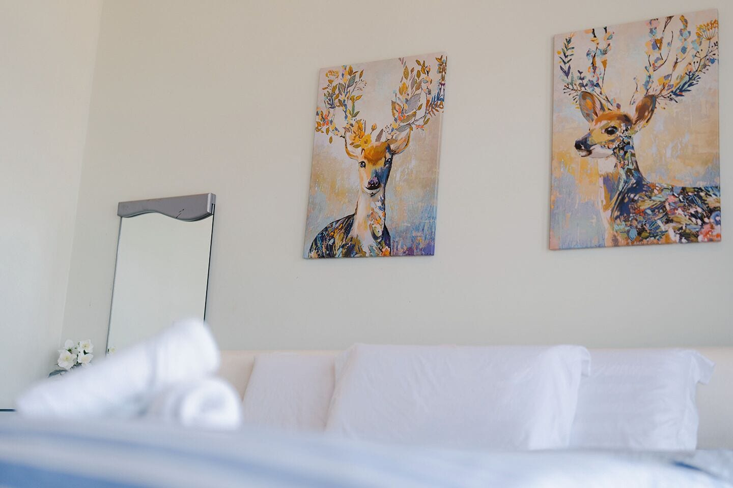 A bedroom with a white bed, a bedside table, and two paintings of deer on the wall.