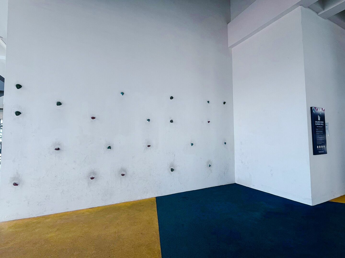 A corner of a room with a white wall featuring a series of evenly spaced holes, a yellow and blue carpet, and a black sign on the adjacent wall.