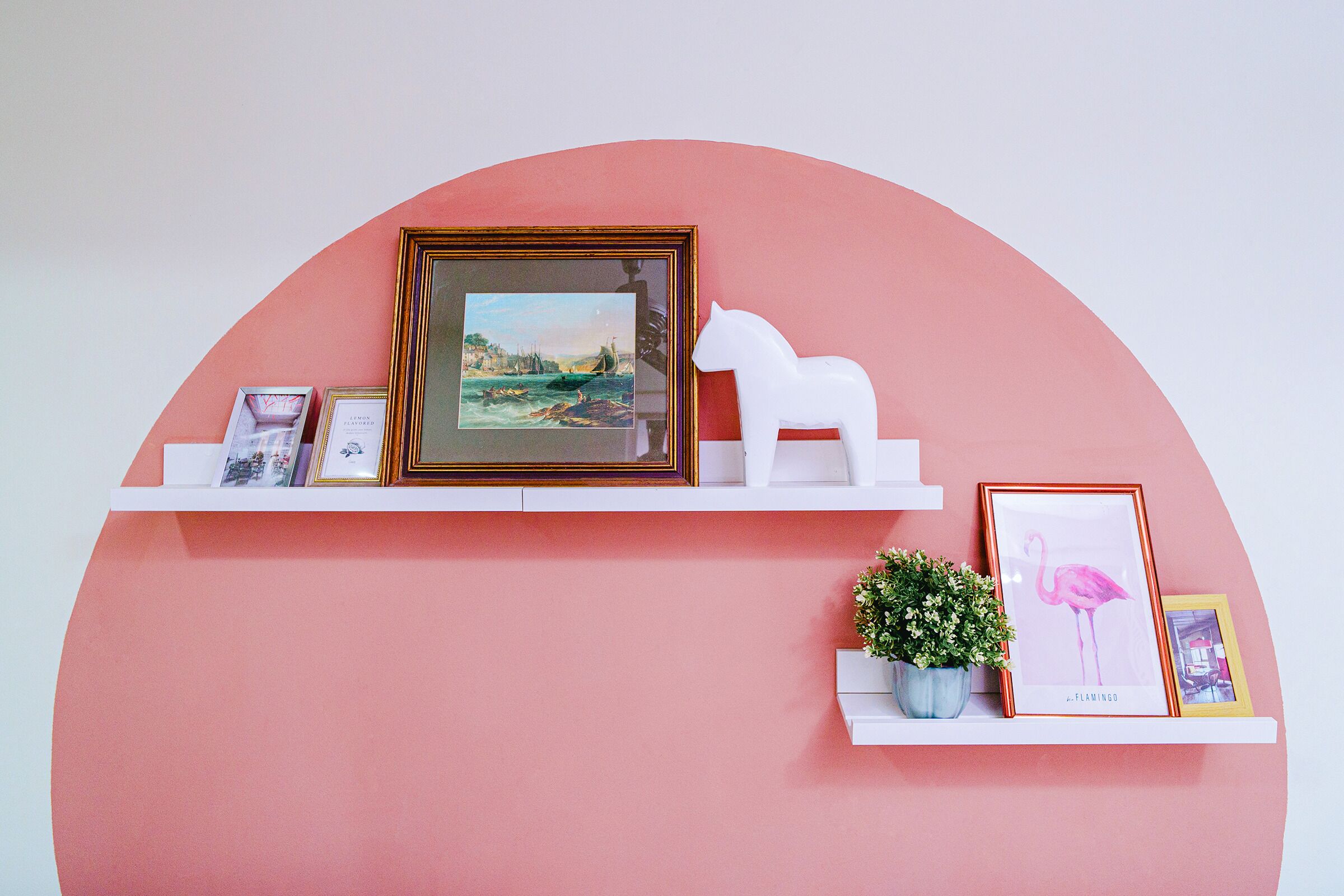 Eine Wand mit einem weißen Regal, das gerahmte Bilder, eine weiße Skulptur und einen Blumentopf zeigt.
