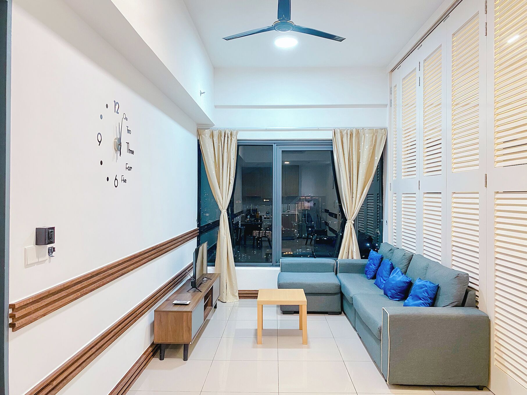A modern living room with a sofa, a wooden side table, a ceiling fan, and a large window with curtains.