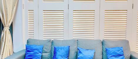 A grey sofa with blue cushions in front of a wall with multiple window blinds.