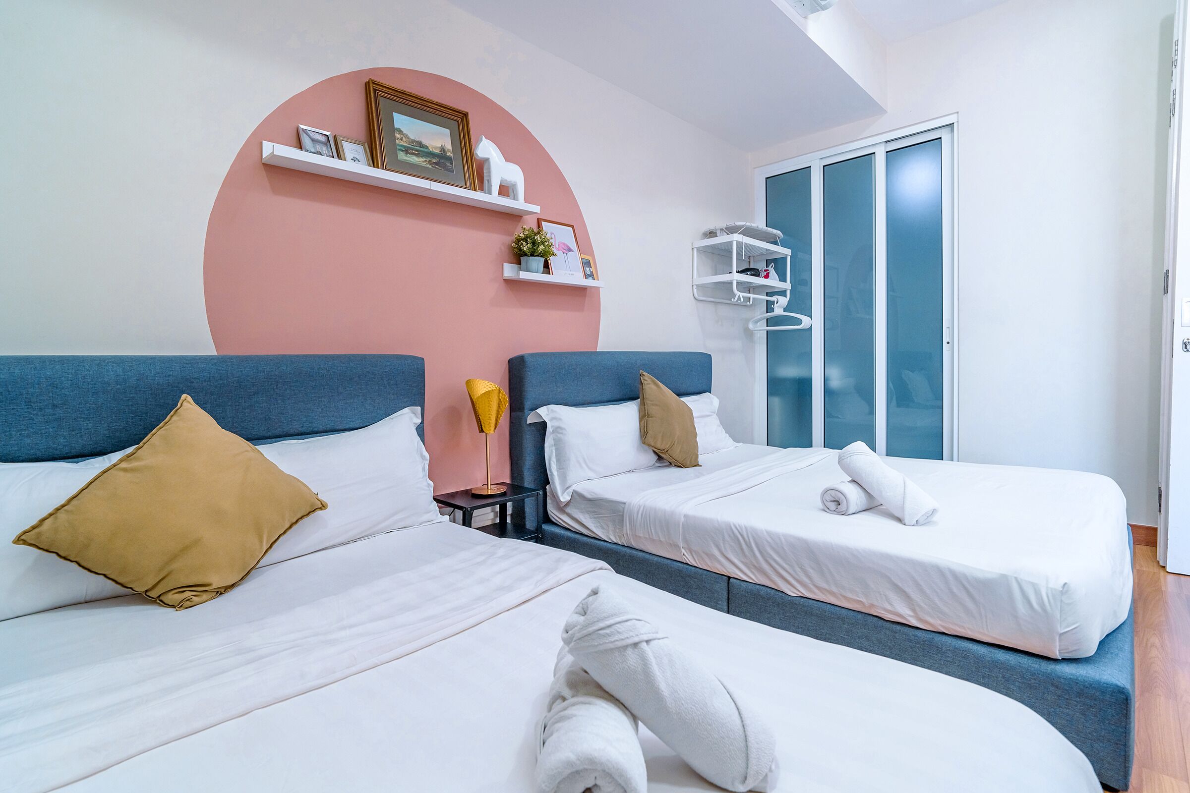 Quarto de hotel com duas camas, uma parede de destaque rosa e uma porta branca com painéis de vidro jateado.