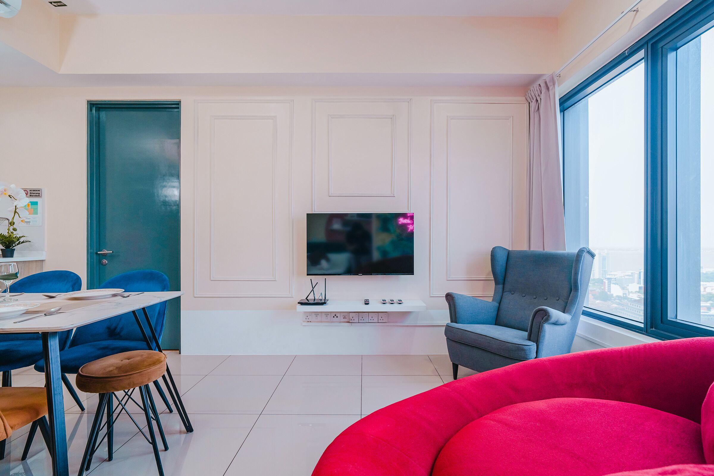 A modern living room with a dining area, a large window with a city view, a flat-screen TV, and a grey armchair.