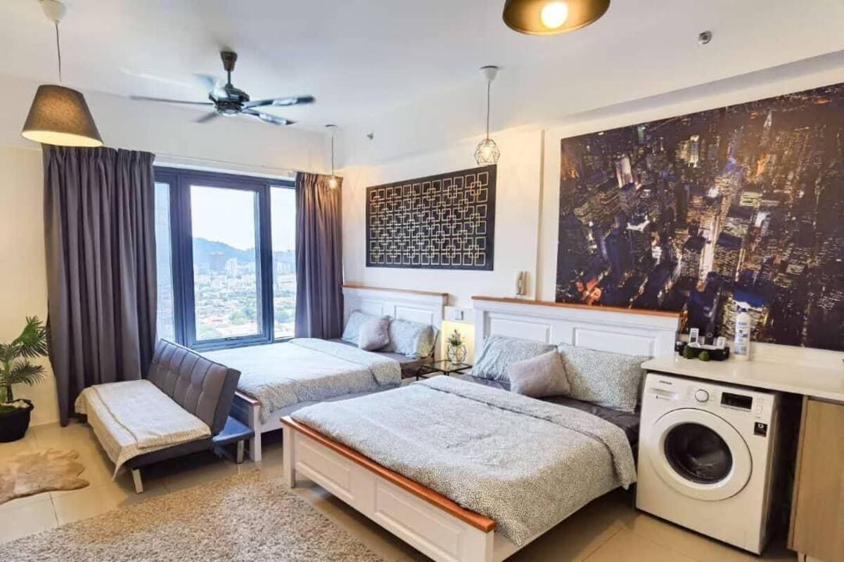 A modern bedroom with two beds, a ceiling fan, a large window with curtains, a cityscape painting, a washing machine, and a small seating area.