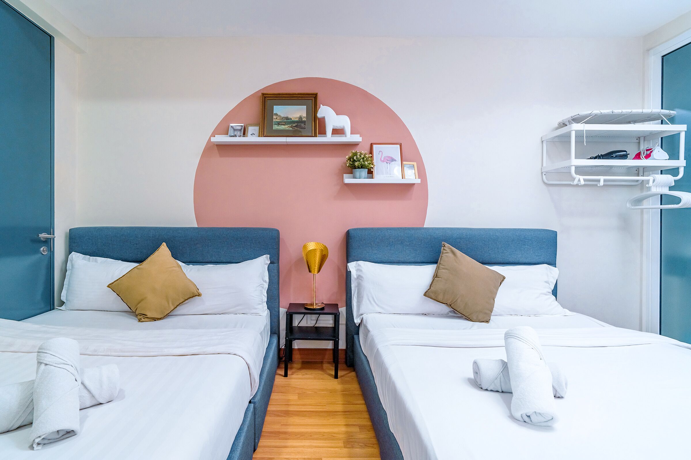 A hotel room with two beds, a pink accent wall, a small table with a lamp, and a white shelf with towels.