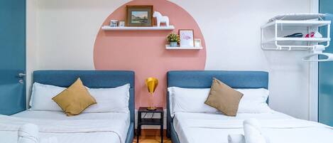 A hotel room with two beds, a pink accent wall, a small table with a lamp, and a white shelf with towels.