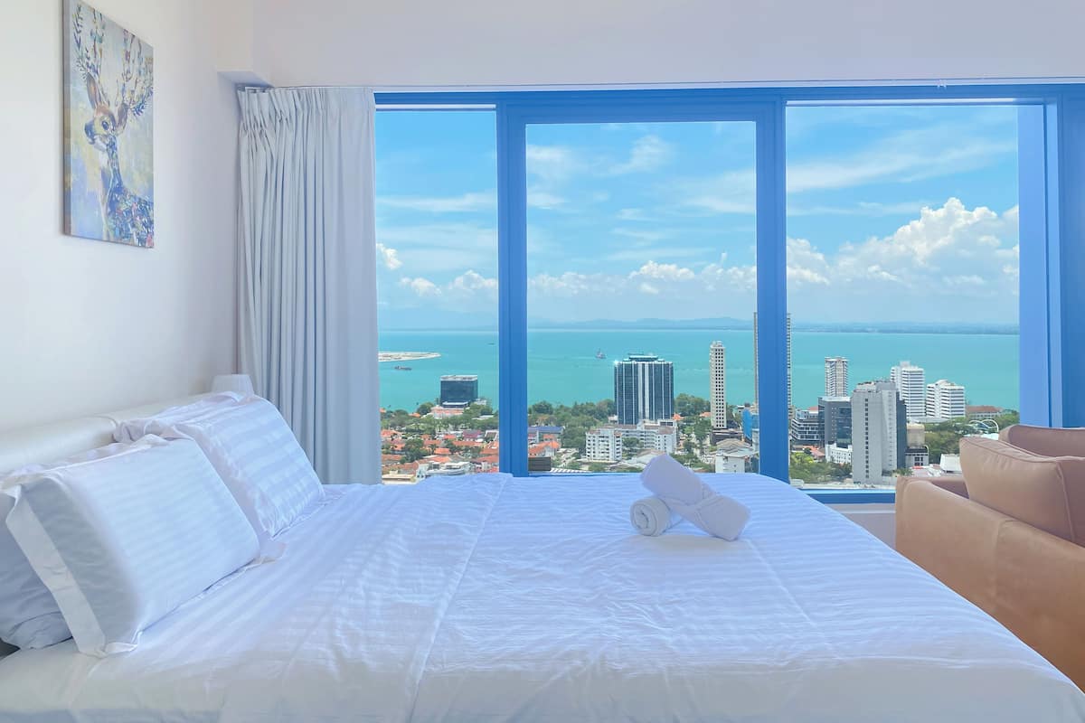 A hotel room with a large bed, a view of the city and sea, and a painting on the wall.