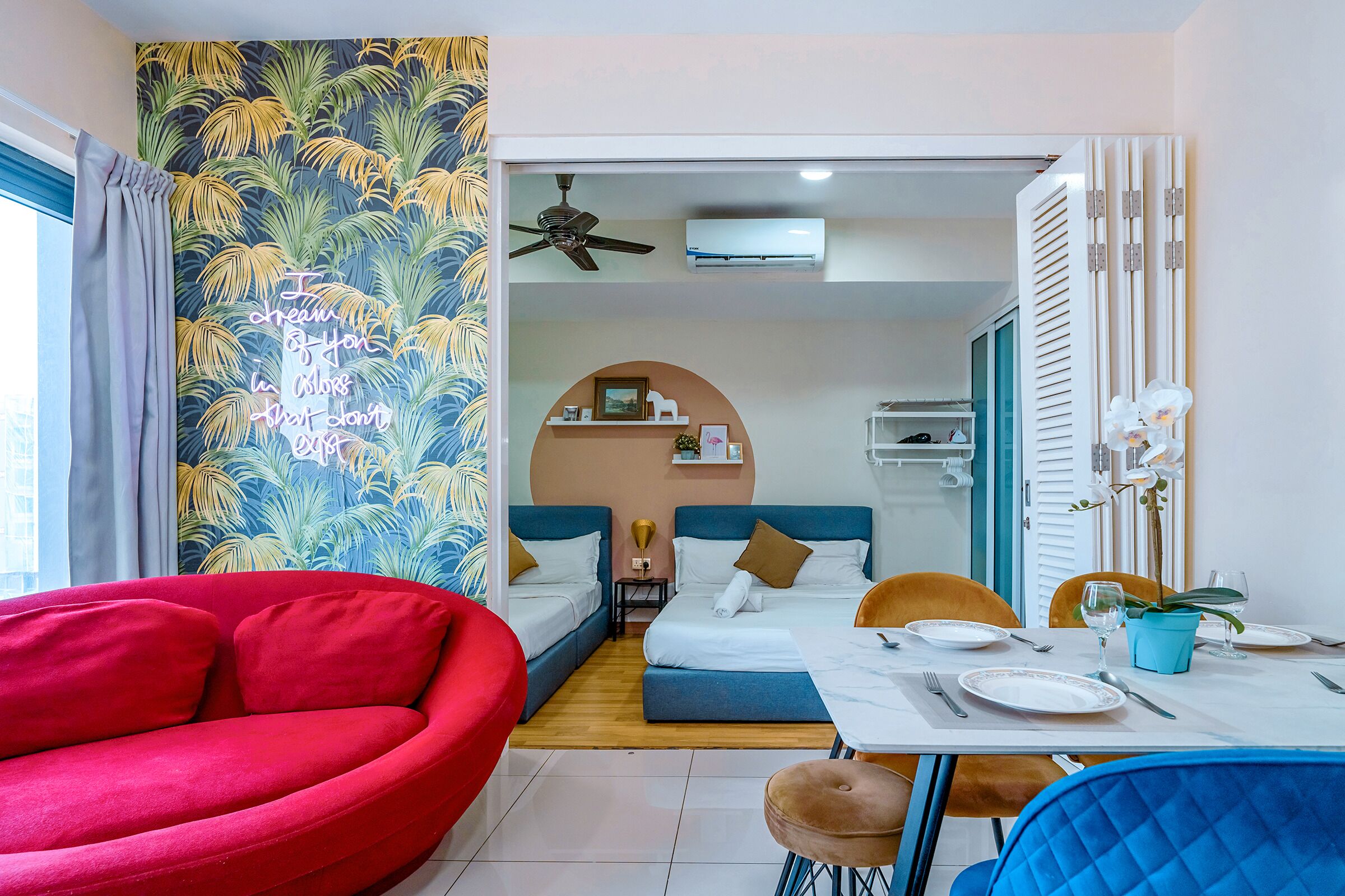 A hotel room with a bed, a red chair, a dining table with plates and cutlery, and a wall with a tropical leaf pattern.