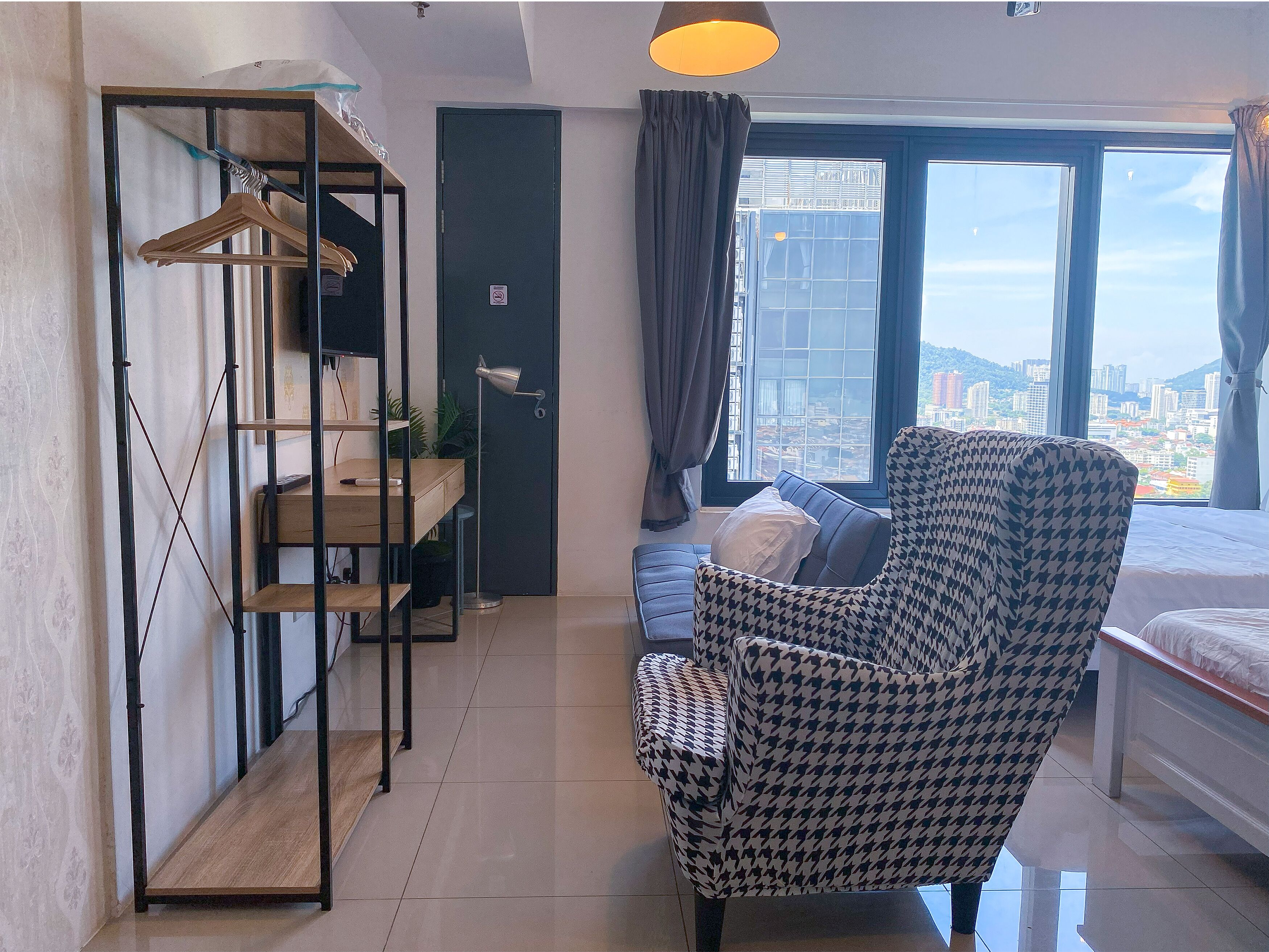 Una habitación de hotel moderna con un ventanal grande, un sillón con estampado de piedritas, un escritorio de madera y una estantería de metal.