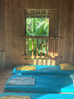 A room with a bamboo wall, a window with a view of green foliage, a bed with blue sheets, and a wooden bench.