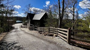A rustic cabin with a gravel driveway and wooden fence, surrounded by trees and a clear sky. - Fels Haus- Hot Tub, WiFi, Gas Grill, located on Cliffview Resort (Campton)