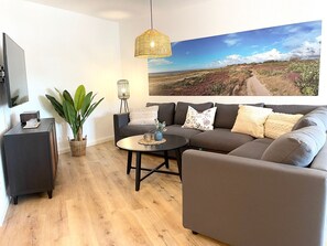Ein modernes Wohnzimmer mit Sofa, Couchtisch, Fernseher, Pflanze und einem großen Landschaftsbild.