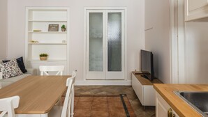 Une cuisine compacte avec un coin repas, des meubles blancs, une table en bois et un téléviseur posé sur un meuble en bois.