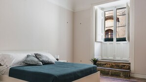 Une chambre à coucher avec un lit, une table de chevet, une fenêtre donnant sur un immeuble et un plancher carrelé.
