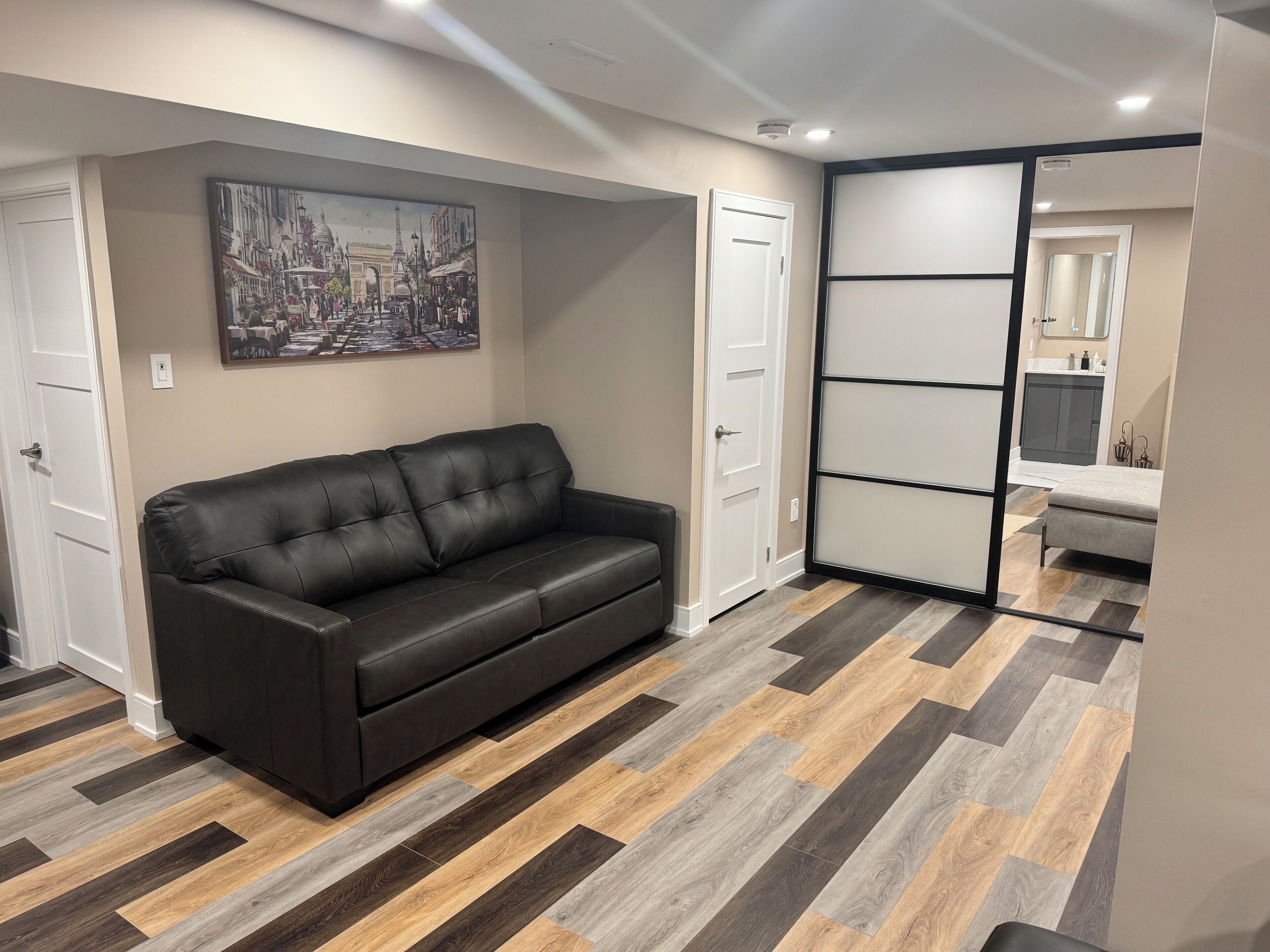 Una sala de estar moderna con un sofá de cuero negro, un cuadro de un paisaje urbano y un suelo con estampado de espiga.