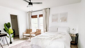 Ein Schlafzimmer mit einem großen Bett, zwei Stühlen, einem Schreibtisch, einem Deckenventilator und einem Fenster mit Vorhängen.