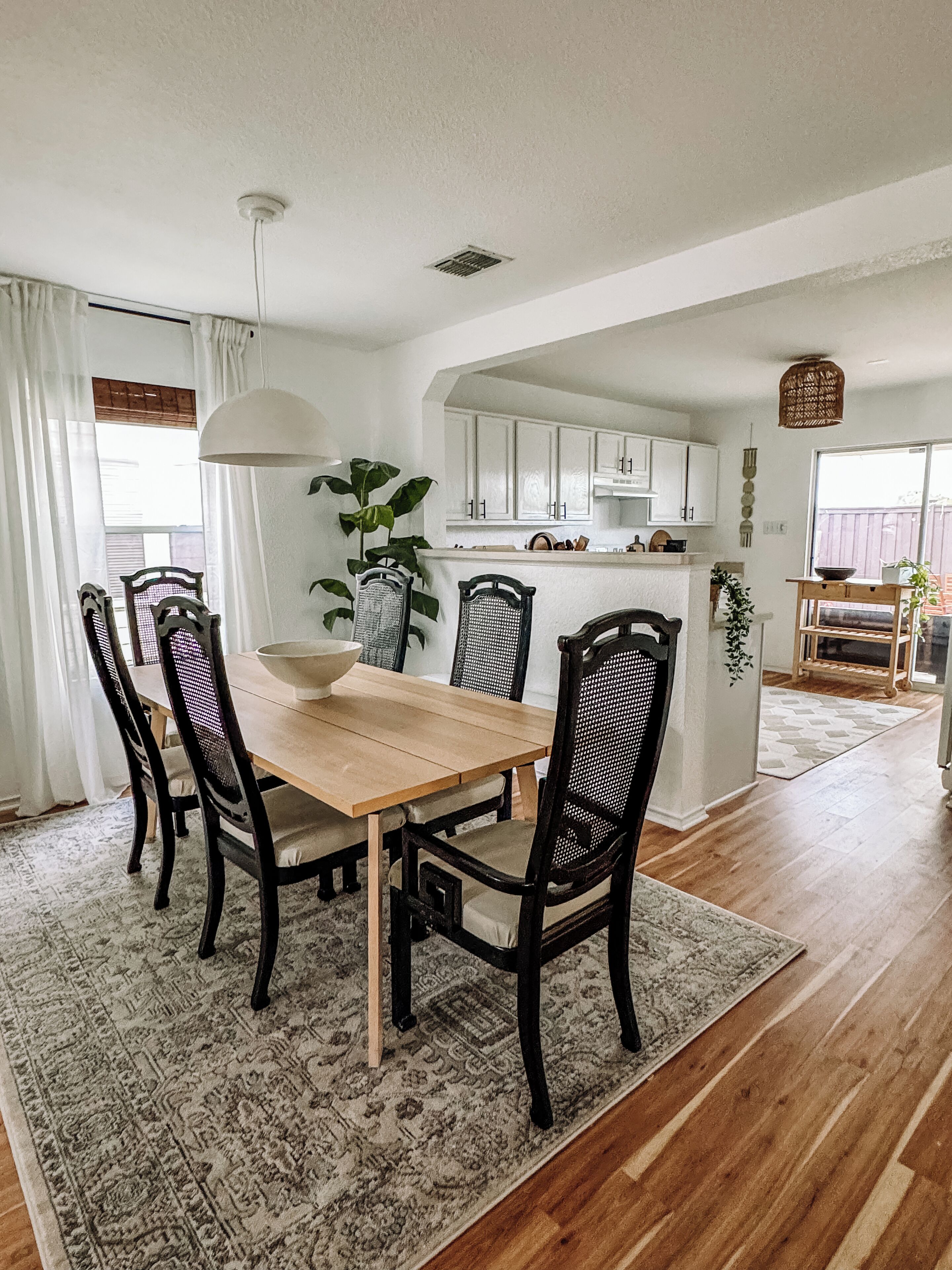 A dining area with a wooden table and four chairs, a rug, and a kitchen in the background.