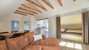 Een moderne loft met zichtbare houten balken, een hoekbank, een bed met gordijnen, een eethoek en een keuken op de achtergrond.