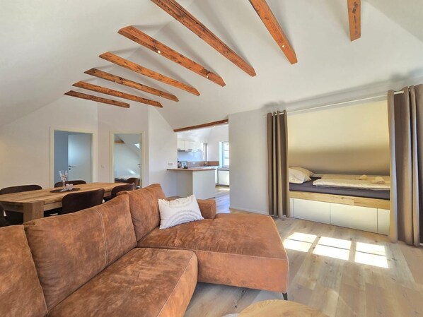 A modern loft-style room with exposed wooden beams, a sectional sofa, a bed with curtains, a dining area, and a kitchen in the background.