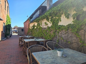 An outdoor seating area with wicker tables and chairs, a brick pathway, and a building with ivy-covered walls.