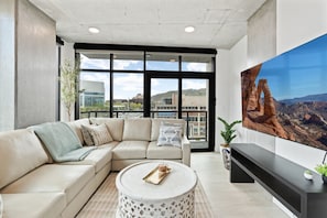 A modern living room with a beige sectional sofa, a round coffee table, and a large flat-screen TV mounted on the wall.