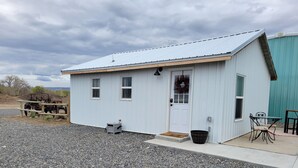 A small, single-story building with a white exterior and a sloped roof. There is a door with a wreath, a small porch area, and a table with chairs outside.