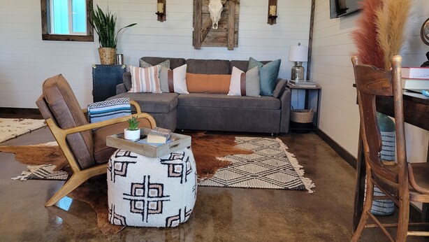 A cozy living room with a sectional sofa, a wooden chair, a patterned ottoman, and a rustic ceiling.
