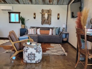 A cozy living room with a sectional sofa, a wooden chair, a patterned ottoman, and a rustic ceiling. - Cozy Guest House at the base of the Big Horn Mountains (Lovell)