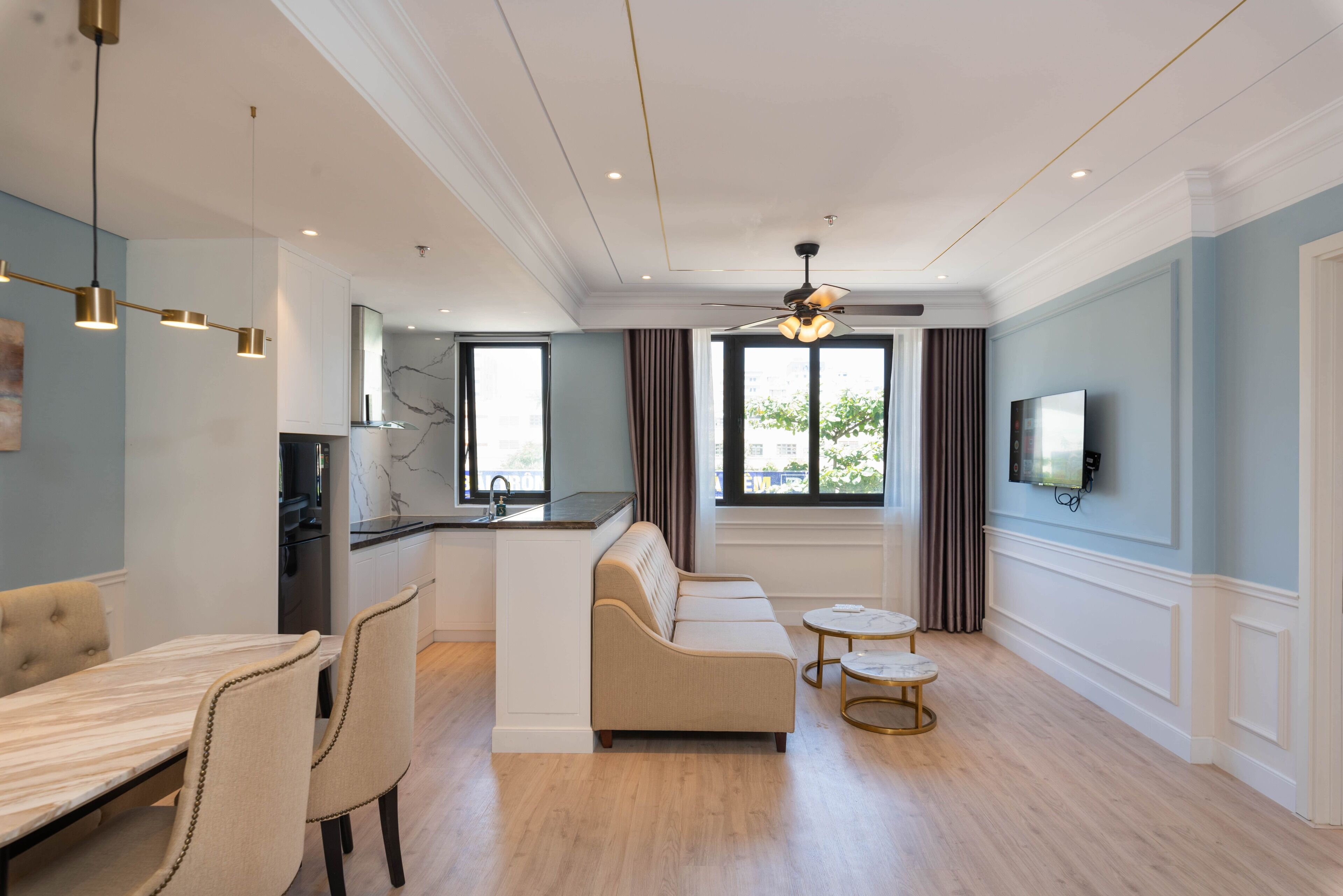 A modern living room with a sofa, dining table, and chairs, a kitchen area, and a ceiling fan.