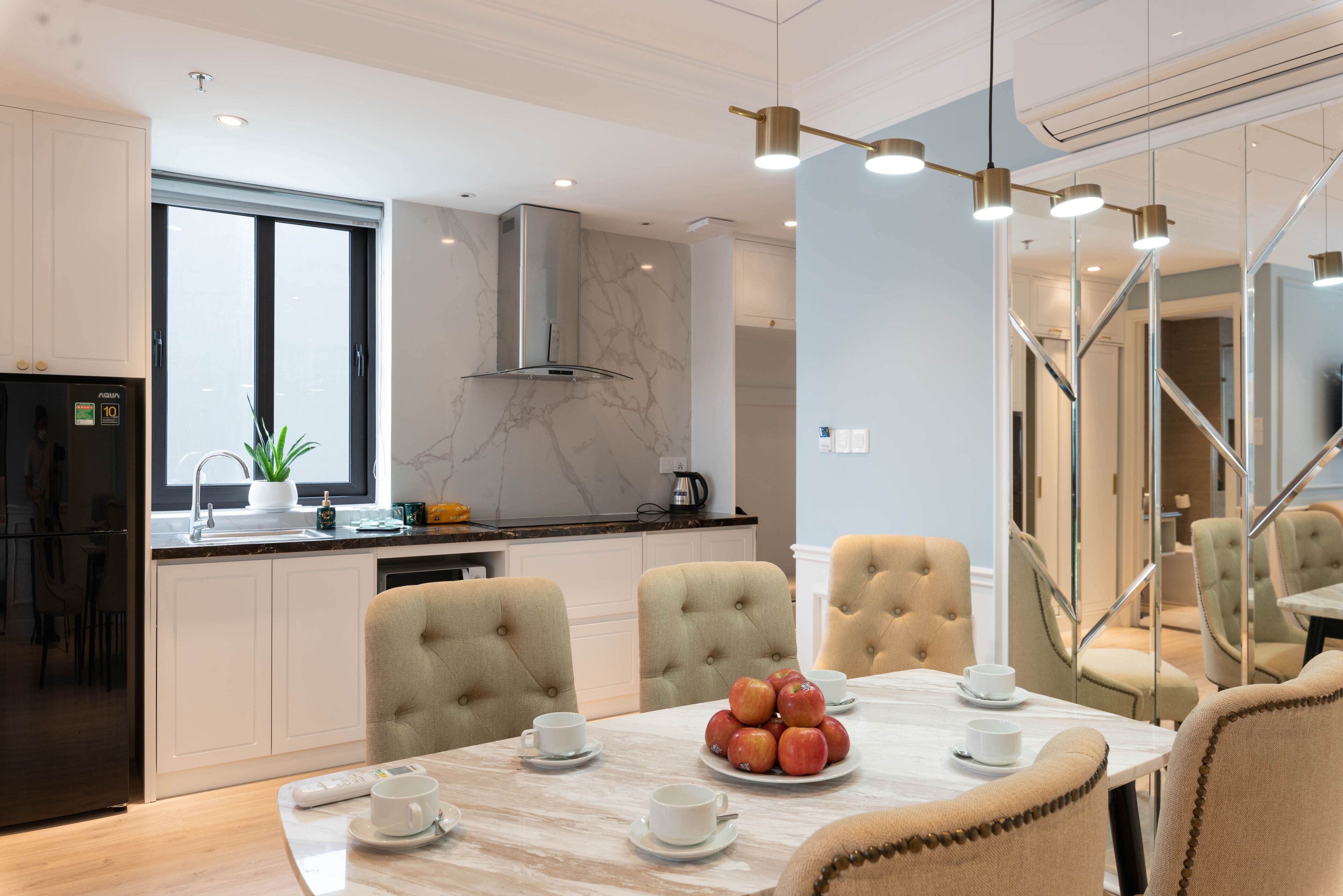 A modern kitchen with white cabinetry, a marble backsplash, and a built-in stove. There is a dining area with a marble table, beige upholstered chairs, and a fruit bowl.