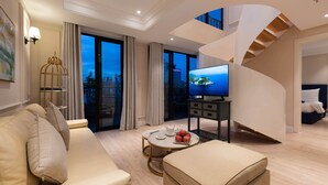 Quarto de hotel moderno com sofá, banquinho, mesa de centro redonda com maçãs, televisão e escada.