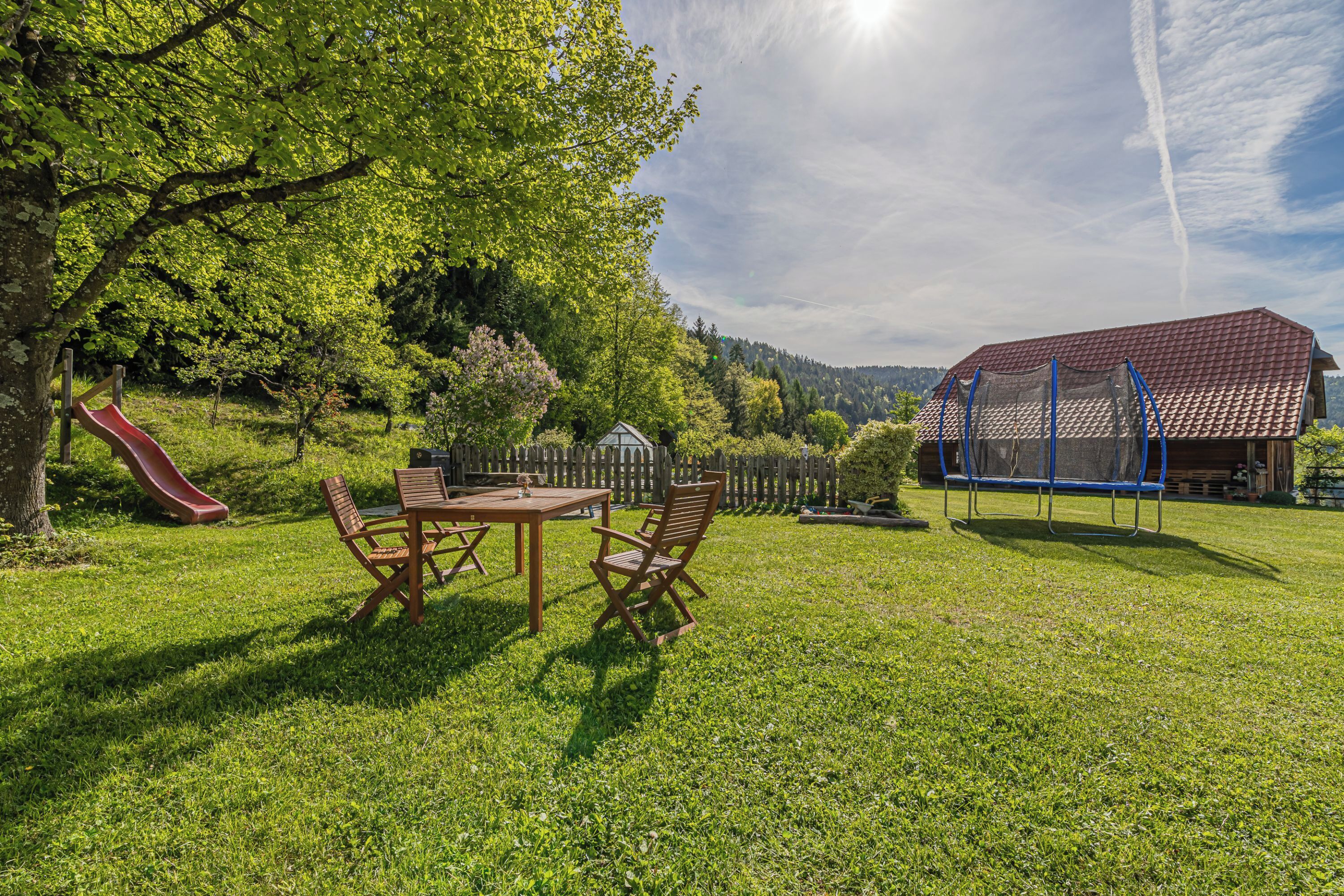 Un espace extérieur verdoyant avec une table de pique-nique et des chaises, un toboggan, une aire de jeux clôturée et un bâtiment avec un toit rouge.