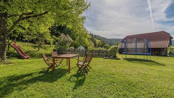 Un espace extérieur verdoyant avec une table de pique-nique et des chaises, un toboggan, une aire de jeux clôturée et un bâtiment avec un toit rouge.