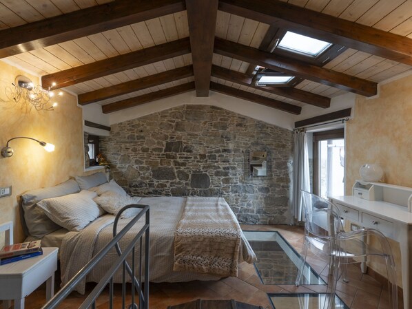 A bedroom with a stone wall, wooden beams, a bed, a nightstand, a lamp, and a window.