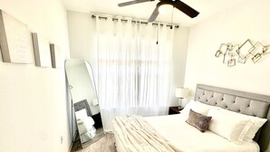 A neatly arranged bedroom with a large bed, a bedside table with a lamp, a ceiling fan, and a window with sheer curtains. - Apartment in the Heart of Frisco (Frisco)