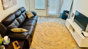 A modern living room with a leather sofa, a woven rug, a flat-screen TV, and a large window with blinds. - Apartment in the Heart of Frisco (Frisco)