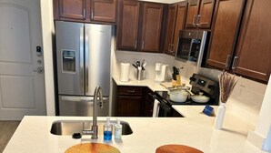 A modern kitchen with stainless steel appliances, wooden cabinets, a white countertop, and two white bar stools. - Apartment in the Heart of Frisco (Frisco)
