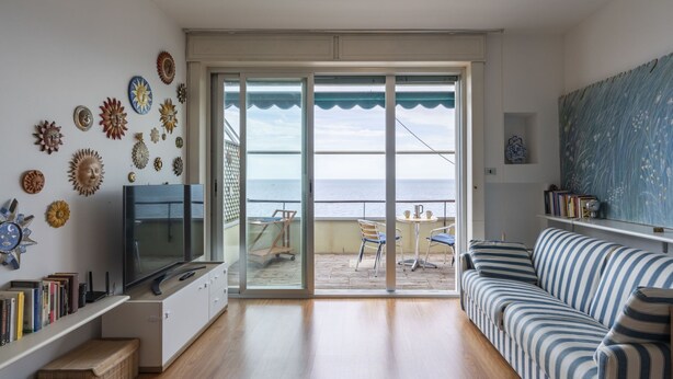 Een woonkamer met een televisie, boekenkasten, een gestreepte bank en uitzicht op de zee door grote glazen deuren.