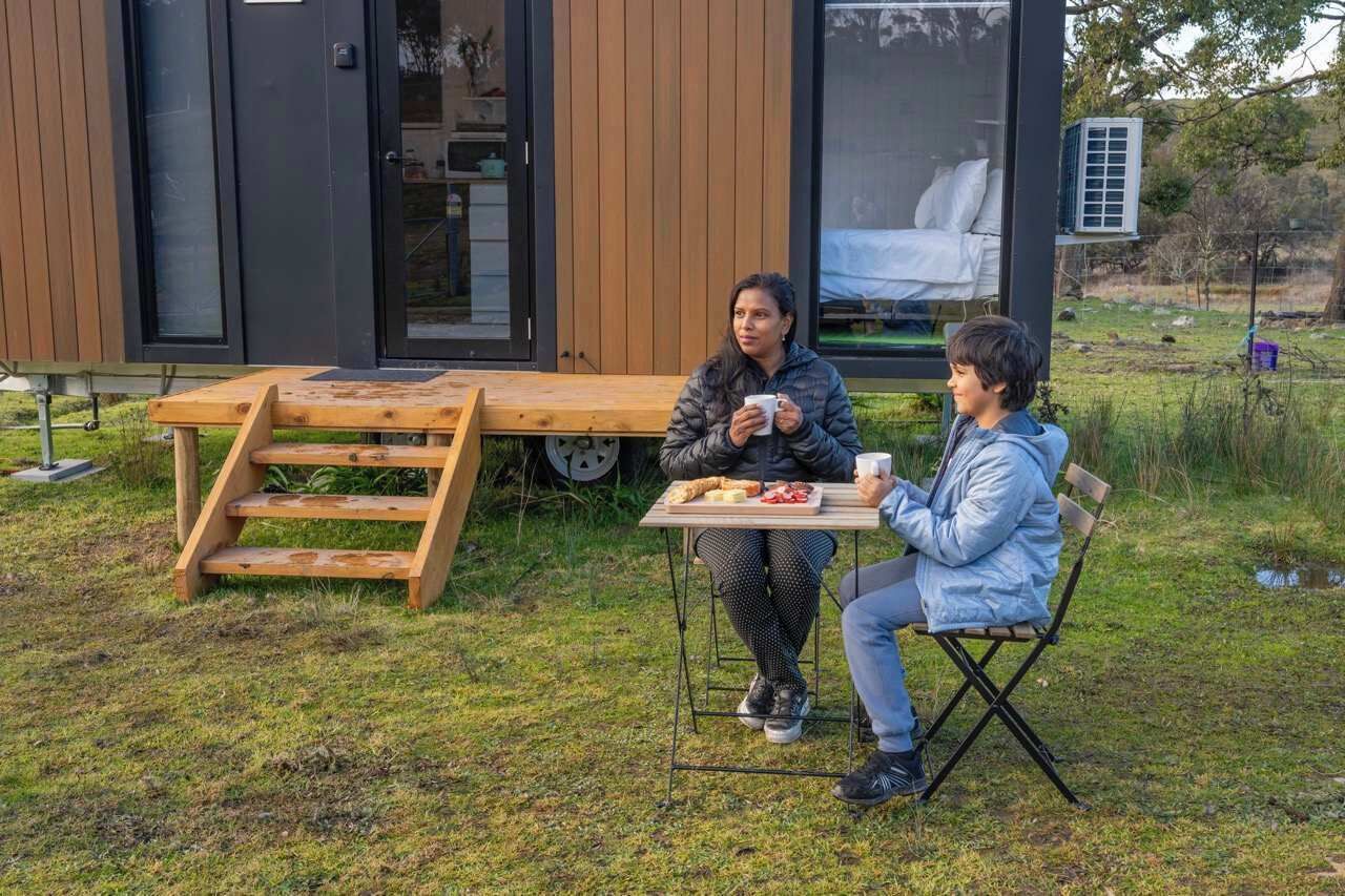 Un espace repas extérieur moderne, avec une table et des bancs en bois, une petite chaise pliante et une vue sur une chambre à coucher équipée d’un lit et d’un climatiseur.