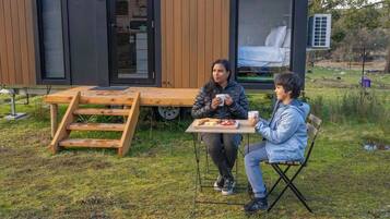 Un espace repas extérieur moderne, avec une table et des bancs en bois, une petite chaise pliante et une vue sur une chambre à coucher équipée d’un lit et d’un climatiseur.