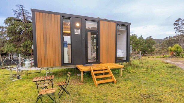 A modern, compact cabin with a wooden exterior, a sliding door, and a small outdoor seating area with a table and chairs. - Sparkling Tiny House by Tiny Away (Windeyer)