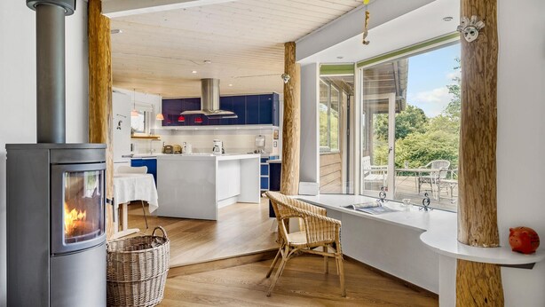A modern kitchen with a wood-burning stove, a dining area with a wicker chair, and a view of the outdoors. - Cozy home in Græsted with sauna (Græsted)