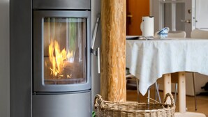 A modern wood-burning stove with a glass door, a wicker basket, and a dining area in the background. - Cozy home in Græsted with sauna (Græsted)