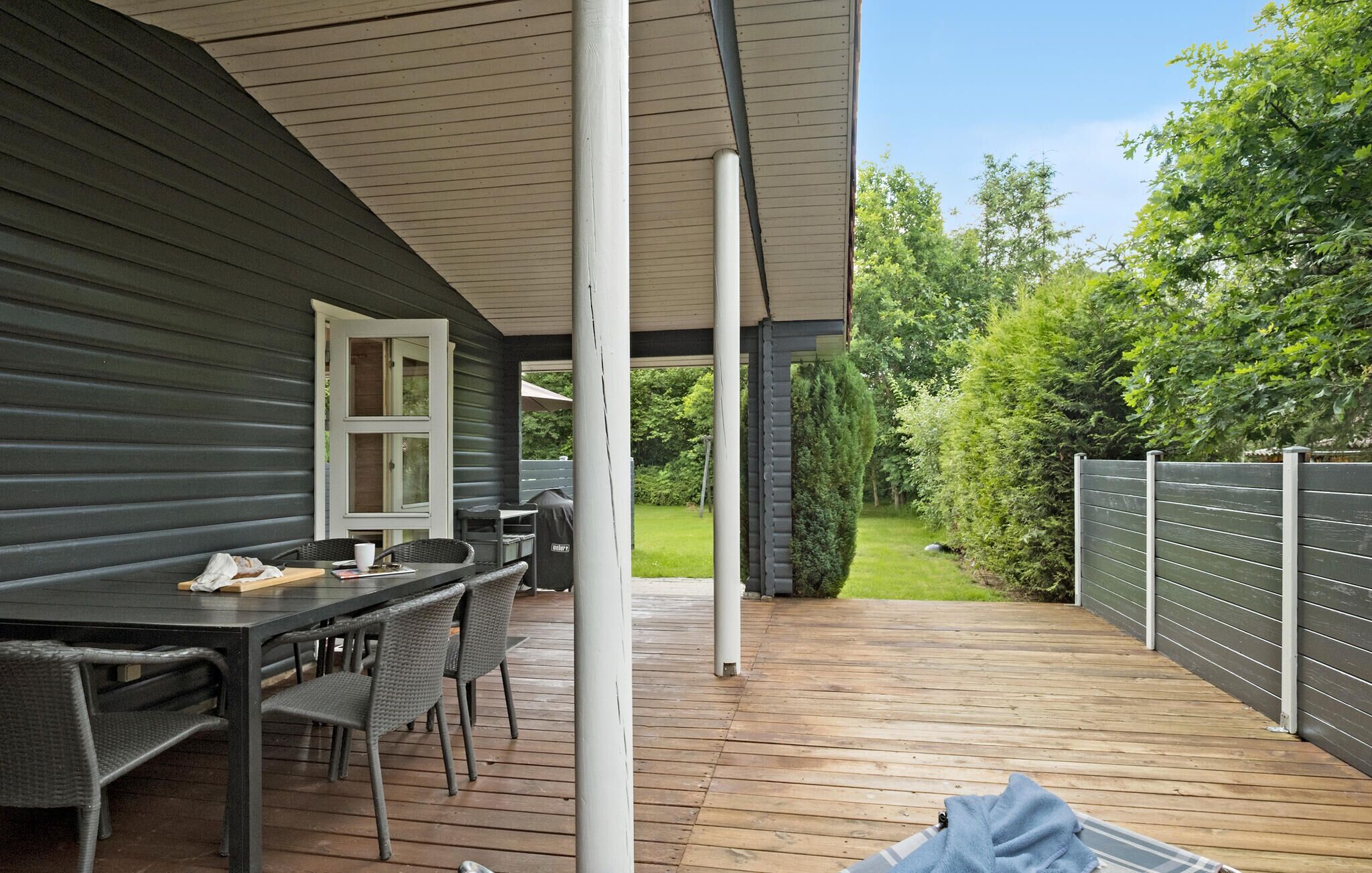 Ein überdachter Patio-Bereich mit Tisch und Stühlen, einem Grill und Blick auf einen grünen Garten.